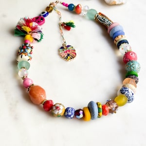 May include: A colourful beaded necklace with a variety of shapes, sizes, and textures. The necklace features a peace sign charm, a leaf charm, and a variety of colourful beads, including glass, ceramic, and wood.