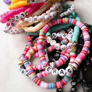 May include: A stack of colorful bracelets with letter beads spelling out words like "LOVE", "ERAS", and "FOLK".