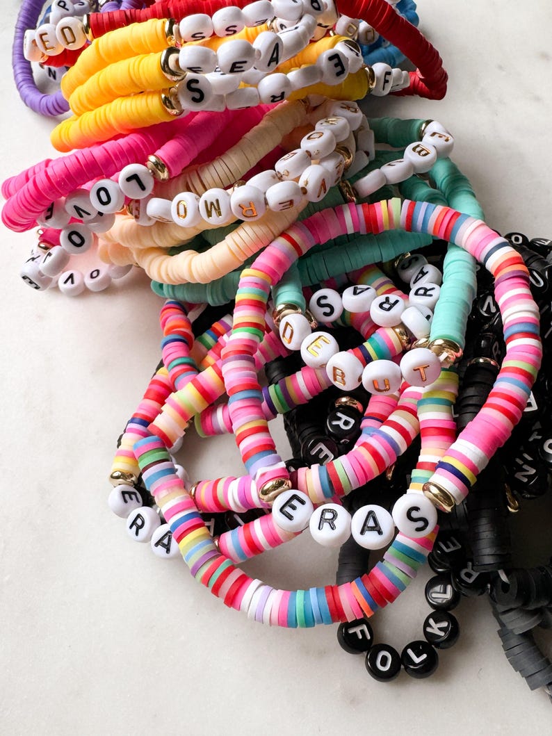 May include: A stack of colorful bracelets with letter beads spelling out words like "LOVE", "ERAS", and "FOLK".