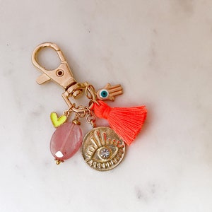 May include: Gold keychain with a pink stone, a yellow heart charm, a blue evil eye charm, a gold hamsa hand charm, and a bright orange tassel.