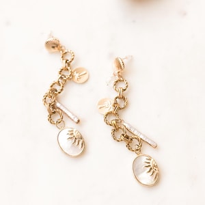 May include: A pair of gold-toned earrings with a nautical theme. Each earring features a chain of linked circles, a small disc, a white oval pendant with a sun design, and a small bar. The earrings are set against a white marble surface.