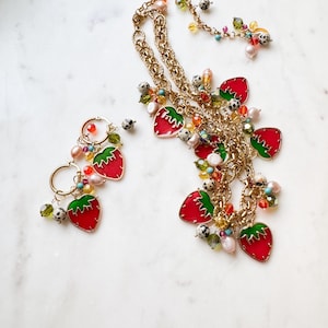 May include: A gold-toned necklace and matching earrings set. The necklace and earrings feature red strawberry charms with green leaves, accented with colorful beads and pearls. The jewelry is set against a white marble background.