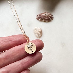 May include: A gold-colored necklace with a round pendant featuring a palm tree design. The necklace is held in a hand, with seashells and driftwood in the background. The pendant is approximately 0.75 inches in diameter.