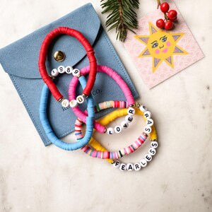 May include: Four colorful beaded bracelets with different words spelled out on them. The bracelets are red, blue, pink, and yellow. The words on the bracelets are "1989", "RED", "LOVER", and "FEARLESS". The bracelets are in a blue velvet pouch with a small card with a sun illustration.