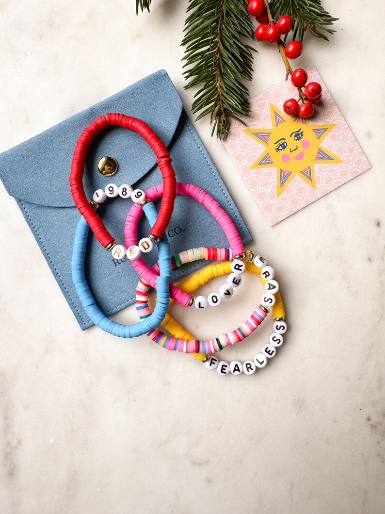 May include: Four colorful beaded bracelets with different words spelled out on them. The bracelets are red, blue, pink, and yellow. The words on the bracelets are "1989", "RED", "LOVER", and "FEARLESS". The bracelets are in a blue velvet pouch with a small card with a sun illustration.