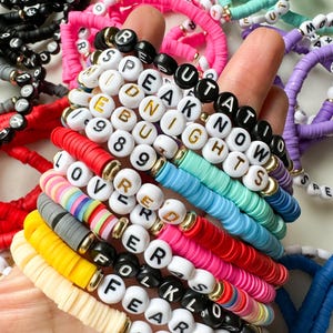 May include: A stack of colorful beaded bracelets with letter beads spelling out words like "Speak Now", "Midnights", "Lover", and "Folklore".