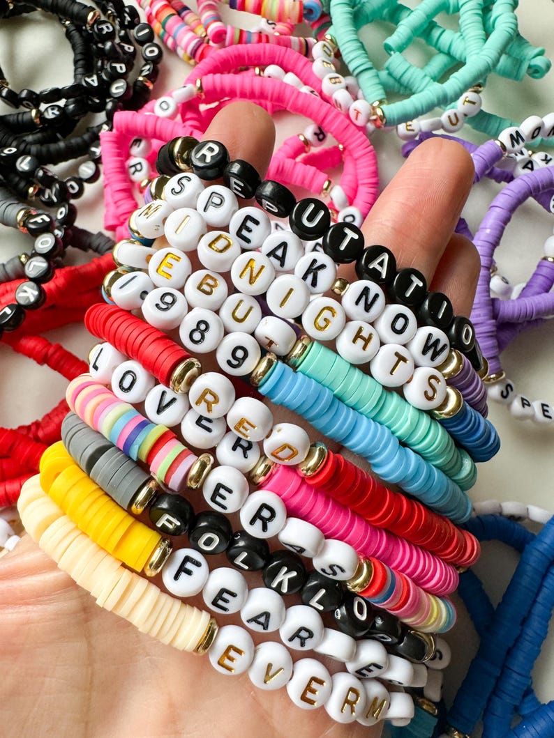 May include: A stack of colorful beaded bracelets with letter beads spelling out words like "Speak Now", "Midnights", "Lover", and "Folklore".