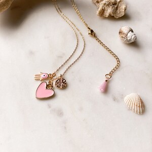 Puede incluir: Un collar dorado con múltiples colgantes, incluyendo un corazón rosa, una mano de Hamsa y un colgante circular. El collar también presenta una pequeña borla rosa. El fondo incluye conchas marinas y un trozo de madera flotante.