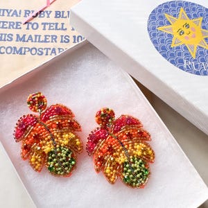 May include: A pair of leaf-shaped beaded earrings in a white box. The earrings feature a gradient of colors, including red, orange, yellow, and green. The box is open, revealing the earrings nestled inside. Pink shredded paper is visible.