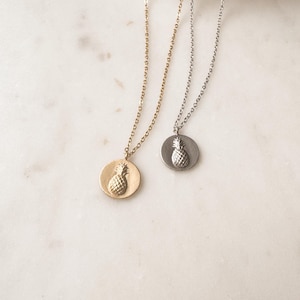 May include: Two gold and silver necklaces with pineapple pendants. The necklaces are on a white marble surface.