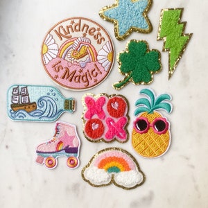 May include: A set of ten colourful iron-on patches featuring various designs, including a rainbow, a pineapple, a roller skate, a bottle with a ship inside, a four-leaf clover, a lightning bolt, a star, and a heart with the text "XO" on it. The patches are all outlined in gold thread and have a fuzzy texture.