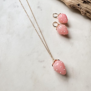 May include: A gold-toned necklace and matching earrings set. The pendant and earrings are shaped like pink strawberries, adorned with small white pearl-like accents. The jewelry is displayed on a white marble surface.