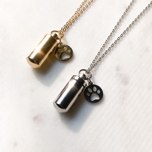 Puede incluir: Dos collares de plata con un pequeño dije cilíndrico y un dije con forma de huella de pata. Uno tiene una cadena dorada y el otro una cadena plateada.