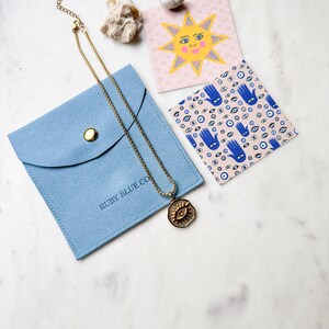 May include: A gold chain necklace with a large gold pendant featuring an evil eye design. The necklace is displayed in a blue velvet pouch with the text "RUBY BLUE CO" printed on it. The pouch is on a white marble surface.