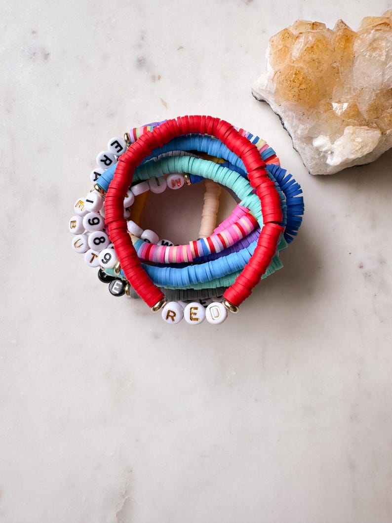 May include: A stack of colorful beaded bracelets with the word "RED" spelled out in white beads with gold lettering.