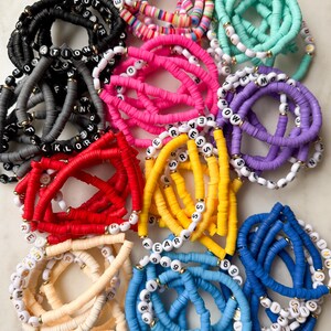 May include: A collection of colorful beaded bracelets with letter and number charms. The bracelets are made of various colors, including red, yellow, blue, green, and pink. The charms are made of gold metal and are spelled out with letters and numbers.