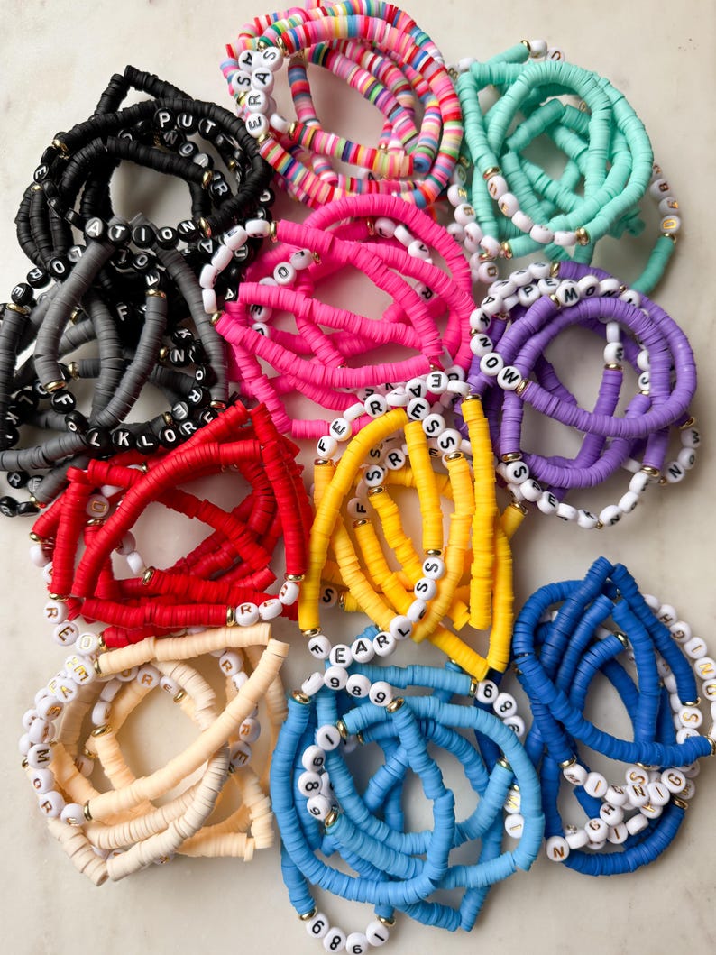 May include: A collection of colorful beaded bracelets with letter and number charms. The bracelets are made of various colors, including red, yellow, blue, green, and pink. The charms are made of gold metal and are spelled out with letters and numbers.