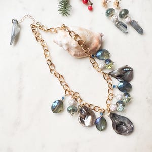 May include: A gold chain necklace with iridescent gray and blue teardrop-shaped pendants and matching earrings. The jewelry is adorned with smaller blue and green beads. A seashell and sprigs of greenery are in the background.