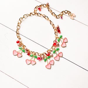 May include: A gold chain necklace with pink and green cherry charms. The necklace features a variety of red, pink, and green beads.