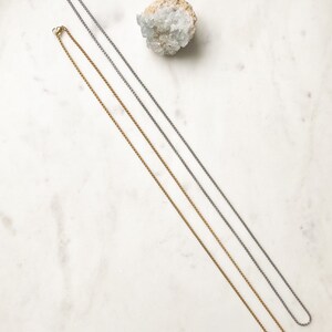 May include: Two delicate chain necklaces, one silver and one gold, lay on a white marble surface.