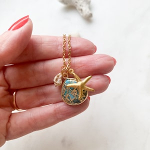 Puede incluir: Collar dorado con un colgante redondo con una piedra turquesa y un dije de estrella de mar dorada. Un pequeño dije de concha cuelga junto al colgante. El collar se sostiene en una mano con esmalte de uñas rojo.