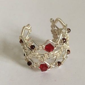 Può includere: Un anello in filo d'argento con perline rosse e marroni. L'anello è realizzato con più strati di filo, creando un effetto intrecciato. Le perline rosse sono sfaccettate e le perline marroni sono rotonde.