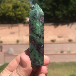 May include: A green and purple gemstone point, also known as a crystal, is held in a hand. The point is smooth and polished, with a pointed tip and a flat base.