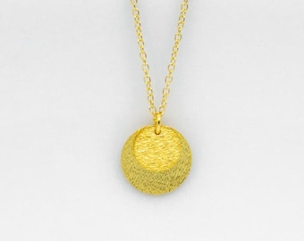 Può includere: Una collana color oro con una catenina sottile e un ciondolo circolare. Il ciondolo presenta una finitura opaca e testurizzata con un design a strati orizzontali, che crea una sottile forma a mezzaluna nella parte superiore del disco.