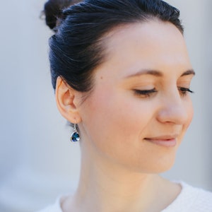May include: A woman with dark hair pulled back in a bun wears a white shirt and silver earrings with a dark blue gemstone.