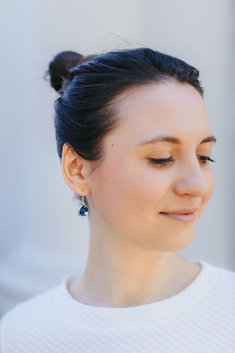 May include: A woman with dark hair pulled back in a bun wears a white shirt and silver earrings with a dark blue gemstone.