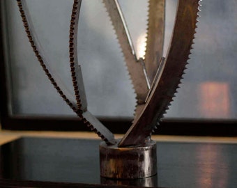May include: A silver metal sculpture of a stylized plant with multiple branches. The sculpture is made from repurposed saw blades and has a textured surface.