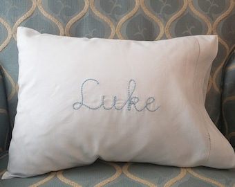 May include: A white pillow with blue embroidery that reads "Luke" on a light blue patterned chair.