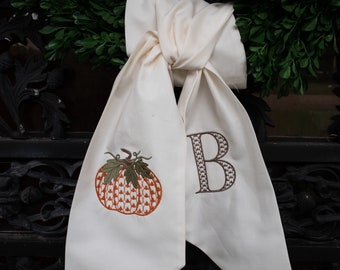 May include: A white fabric bow with an embroidered pumpkin and the letter "B" in brown thread. The bow is tied around a wreath.