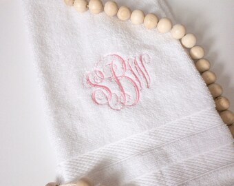Può includere: Asciugamano bianco con monogramma rosa delle lettere SBW. L'asciugamano ha un bordo strutturato ed è circondato da una collana di perline di legno. L'asciugamano è piegato e posto su una superficie bianca.