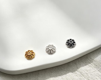 May include: Three small flower-shaped stud earrings in gold, silver, and black. The earrings are displayed on a white, oval-shaped dish. The earrings have a detailed, openwork design.