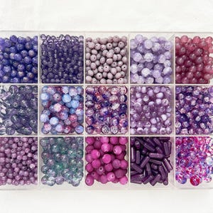 Puede incluir: Una caja organizadora de plástico transparente con quince compartimentos separados llenos de un surtido de cuentas de color púrpura, violeta y magenta. La colección incluye varias formas, tamaños y texturas, desde esferas redondas lisas hasta cuentas cilíndricas.