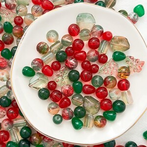 May include: A close-up of a white plate filled with red and green glass beads. The beads are various shapes and sizes, including round, oval, and teardrop.
