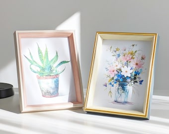 May include: Two picture frames with different colored frames and artwork. The frame on the left is light pink with a white mat and a watercolor painting of a green succulent plant in a pot. The frame on the right is light blue with a gold trim and a watercolor painting of a bouquet of pink, white, and blue flowers in a glass jar.