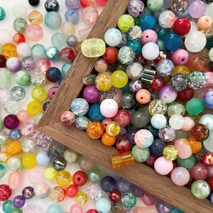 Op de afbeelding: Een selectie van kleurrijke glazen kralen in een houten doos. De kralen zijn in verschillende vormen en maten, waaronder rond, vierkant en ovaal. De kralen zijn een mix van heldere, ondoorzichtige en iriserende kleuren.