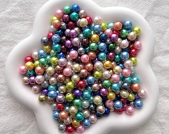 May include: A large assortment of colorful iridescent pearl beads in various shades of pink, blue, green, and purple. The beads are arranged in a star-shaped design on a white ceramic plate.