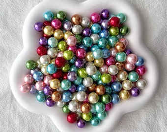 May include: A large assortment of colorful pearl beads in a white flower-shaped dish. The beads are iridescent and come in a variety of colors, including pink, blue, green, and purple.