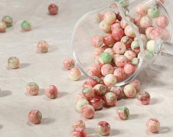 May include: A glass bowl filled with pink and green patterned beads spills onto a white surface. The beads are round and have a glossy finish.