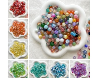 May include: Assortment of colourful glass beads in various shapes and sizes, displayed in a white dish with a flower-shaped design. The beads are arranged in a circular pattern, with the centre of the dish containing a larger cluster of beads.