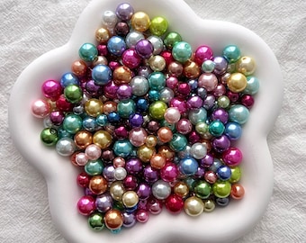 May include: A white ceramic bowl shaped like a flower with a large assortment of iridescent pearl beads in various colors, including pink, blue, green, and purple.