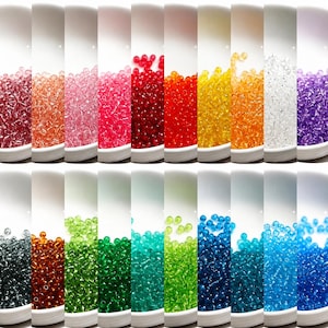May include: A collection of small, round, translucent glass seed beads displayed in twenty separate white containers. The beads are arranged in a spectrum of colours including red, pink, orange, yellow, green, blue, purple, clear, and black.