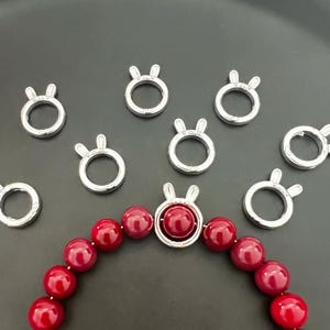 Op de afbeelding: Zilverkleurige metalen sluitingen met konijnenoordetails, gerangschikt rond een rode kralen armband.