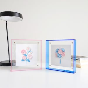 Könnte beinhalten: Zwei quadratische Acrylrahmen, einer rosa und einer blau, jeweils mit floralen Kunstwerken. Der rosafarbene Rahmen zeigt ein rosa-weißes Blumenarrangement, der blaue einen blau-rosa Blumenstrauß. Beide stehen auf einer weißen Oberfläche.