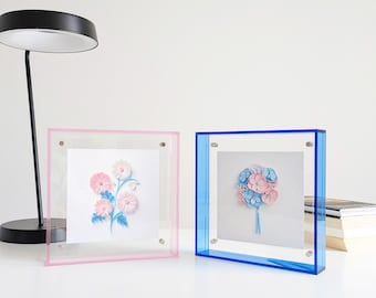 Puede incluir: Dos marcos cuadrados de acrílico, uno rosa y otro azul, cada uno con una obra floral. El marco rosa muestra un arreglo floral rosa y blanco, el azul un ramo azul y rosa. Ambos sobre una superficie blanca, para decoración.