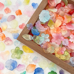 May include: A selection of colourful, translucent flower and leaf-shaped beads in a wooden tray and scattered on a white surface. The beads are in shades of pink, orange, yellow, green, blue, and purple, ideal for crafting or jewellery making.