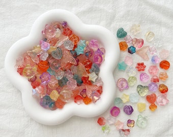 May include: A white cloud-shaped dish filled with colorful translucent plastic beads in various shapes, including stars, flowers, hearts, and wings. The beads are scattered around the dish on a white surface.
